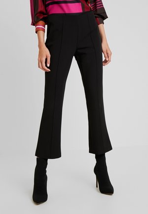 Mujer con pantalones negros de tiro alto y corte cropped con costuras delanteras y botines calcetín negros con tacón sobre fondo liso.