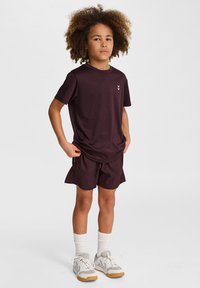 Burgundowy zestaw sportowy składający się z koszulki z krótkim rękawem z małym logo oraz dopasowanych szortów. W komplecie białe skarpetki i jasnoszare buty.