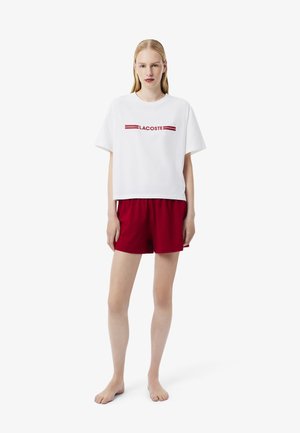 White cotton t-shirt with red "LACOSTE" logo, paired with red shorts. The t-shirt has a relaxed fit and short sleeves. Shorts are loose-fitting.