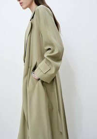 Dlouhý, olivově zelený kabát vyrobený z měkkého materiálu, který má límec, kapsy a mírně oversized rukávy, se hladkými texturami a čistými liniemi.