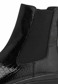 Bottine noire en cuir verni à motif crocodile brillant, avec des panneaux latéraux élastiques et une semelle robuste. Présente une finition lisse et texturée.