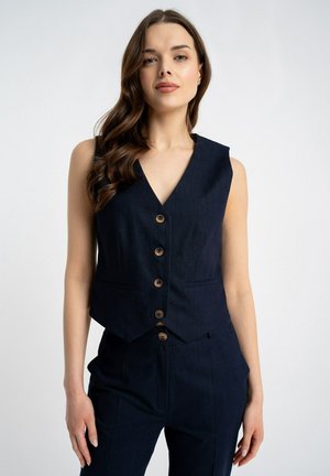 Kobieta z długimi, falującymi włosami ubrana w bezrękawnik z guzikami w kolorze granatowym oraz dopasowane spodnie, stoi na tle jednolitego jasnego tła.