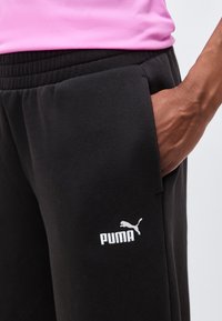 Χέρι ακουμπισμένο στην τσέπη μαύρου παντελονιού φόρμας Puma με ελαστική μέση, άτομο που φοράει ροζ τοπ.