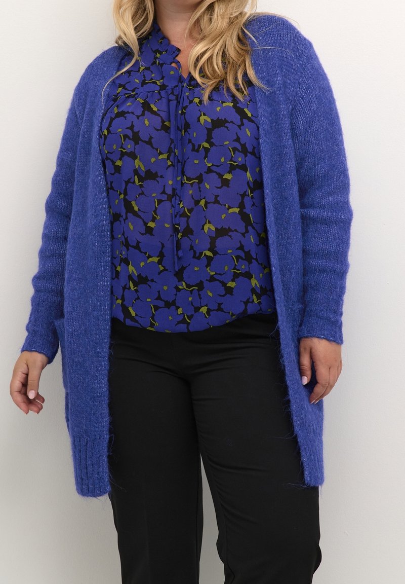 Langer, offen geschnittener Cardigan aus weichem, flauschigem blauen Strick. Darunter eine gemusterte Bluse in Blau und Schwarz mit grünen Akzenten. Schwarze Hosen runden das Outfit ab.