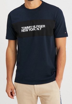Homme portant un t-shirt Tommy Hilfiger bleu marine avec une bande noire et le texte blanc « TOMMY HILFIGER NEW YORK. N.Y », assorti à un pantalon beige.