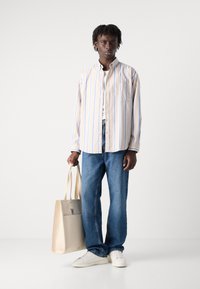 Gestreepte button-up shirt in beige met blauwe lijnen, gecombineerd met blauwe jeans en witte sneakers. Houdt een beige tote bag vast met een vlak ontwerp.