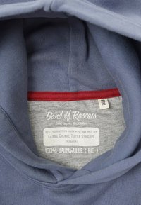 Sininen orgaanisen puuvillan huppari, jossa on harmaa sisäpuoli, punainen reuna sekä hoito-ohjeet ja standardit näyttävä etiketti.