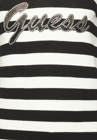 Pull rayé noir et blanc avec un logo "Guess" orné de strass au centre. Tissu en tricot doux avec une texture côtelée.
