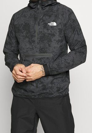 Mężczyzna ubrany w czarno-szarą, wzorzystą bluzę North Face z kapturem i kieszenią z przodu na zamek, z rękami złożonymi na wysokości talii.