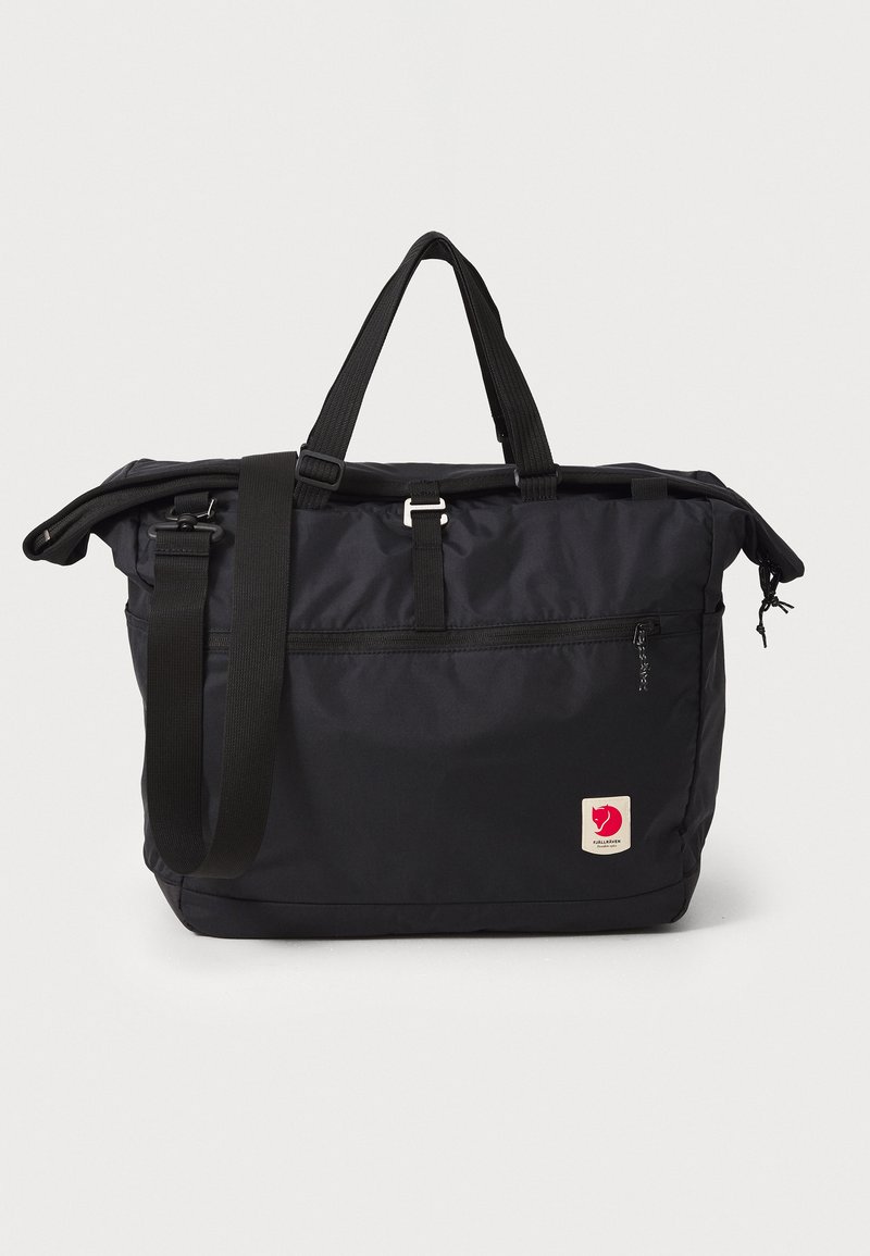Black fabric tote bag with adjustable shoulder strap, top handles, zipper closure, front pocket, and logo patch. Smooth texture, rectangular shape.
