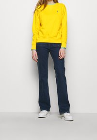 Gul sweatshirt med långa ärmar och blå ärmslut, kombinerad med mörkblå utsvängda jeans och vita sneakers. Har logotyp på bröstet.