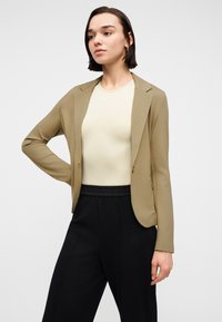 Jeune femme aux cheveux courts et foncés, portant un blazer beige, un haut clair et un pantalon noir, se tenant debout avec une main sur la hanche.