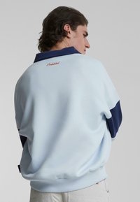 Lyseblå genser med marineblå detaljer, ribbestrikkede mansjetter og nederkant, med brodert "Forbudt"-merke på øvre rygg. Glatt tekstur.
