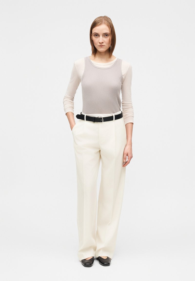 Débardeur gris clair côtelé superposé à un haut à manches longues beige, associé à un pantalon large crème et une ceinture noire. Chaussures noires.