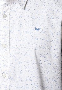 Chemise à motif floral bleu clair en tissu doux. Elle présente un design à boutons et un petit emblème brodé sur la poitrine.
