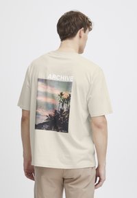 Beige bomull T-skjorte med en rygggrafikk av palmer og en solnedgang, med teksten "ARCHIVE" i hvitt over og "SOLID" under.
