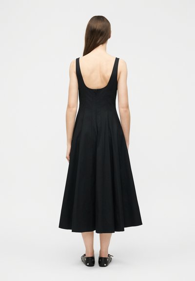 Γυναίκα με μακριά καστανά μαλλιά φορώντας μαύρο μίντι φόρεμα χωρίς μανίκια με χαμηλή πλάτη και μαύρες μπαλαρίνες, στέκεται μπροστά σε απλό λευκό φόντο.