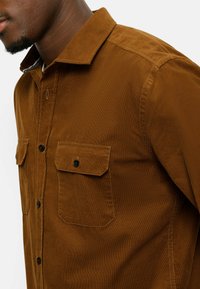 Braunes Cordhemd mit Knopfleiste, zwei Brusttaschen und schwarzen Knöpfen; glatte Textur mit Kragen und subtilen Streifendetails innen.