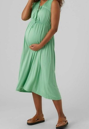 Miętowa zielona sukienka ciążowa bez rękawów z marszczoną talią i detalami na guziki, w zestawieniu z brązowymi sandałami i gładką fakturą.