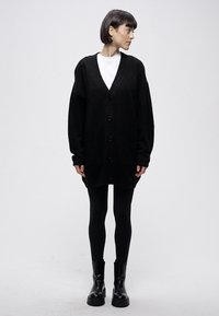 Črni prevelik kardigan z v-izrezom, dolgimi rokavi, zapiranjem na gumbe spredaj in žepi, kombiniran s črnimi legicami in gležnjarji.