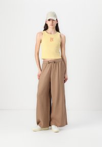 Sárga, rövidített tanktop nyomott mintával, bő szabású barna vászonnadrággal párosítva. A modellen világos sapka és fehér sportcipő található.