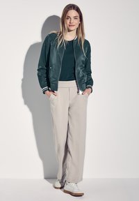 Veste bomber verte avec des accents blancs, associée à un pantalon large beige et des baskets blanches. Texture lisse, design décontracté.