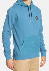 Modrá mikina s kapucí vyrobená z měkké tkaniny, s texturovanou manšestrovou přední kapsou. Má stahovací kapuci a nášivku s logem na hrudi.