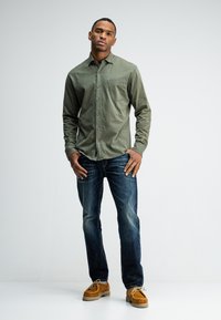 Olijfgroene overhemd met knopen en een borstzak, gecombineerd met donkerblauwe jeans en bruine suède schoenen. De outfit heeft een casual pasvorm.