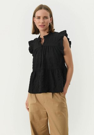 Top texturat negru cu mâneci volănaș, decolteu tip cheie și design în straturi, asociat cu pantaloni bej deschis, pe un fundal neutru.
