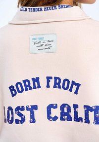 Svetloružová košeľa s modrým textom "BORN FROM LOST CALM" s kvetinovými vzormi a nášivkou s nápisom "NEZABUDNI zamilovať sa do pomalých chvíľ."