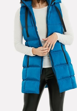 Vrouw draagt een felblauw mouwloos gewatteerd vest over een wit shirt met lange mouwen en zwarte broek, houdt haar handen bij de taille.