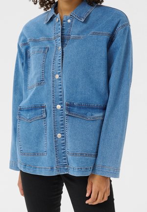 Person iført en blå denimjakke med sølvknapper og tre forlommer, kombineret med sorte bukser mod en ensfarvet baggrund.