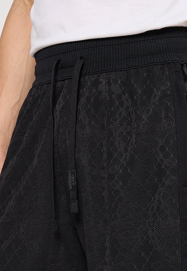 Shorts en maille noire avec taille élastique et cordon de serrage portés avec une chemise blanche, révélant la taille et la partie supérieure des hanches.