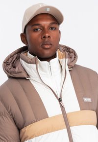 Chaqueta acolchada con una combinación de colores blanco y marrón, con una franja beige, cuello alto, cierre de cremallera y capucha. Llevada con una gorra beige.