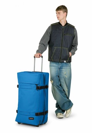 Valise bleue à roulettes avec accents noirs, poignée supérieure et boucles latérales. Tissu texturé et roues lisses pour une mobilité facile.