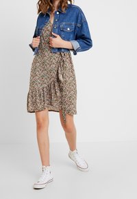 Jeansowa kurtka noszona na zielonej sukience w kwiaty z falbanami, z krótkimi rękawami i bocznym wiązaniem. Białe trampki na wysokiej podeszwie dopełniają stylizację.