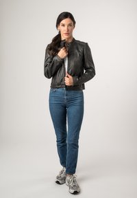 Schwarze Lederjacke mit rundem Kragen, Reißverschluss vorne und langen Ärmeln. Getragen über einem weißen Oberteil mit blauen Jeans und Sneakers.