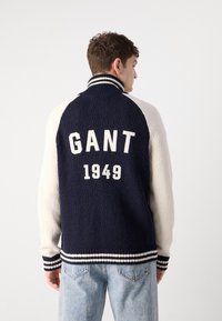 Námornícky a krémový fleecový kabát s vysokým golierom, pruhovanými manžetami a veľkým grafickým nápisom "GANT 1949" na chrbte. Textúrovaná látka so športovým dizajnom.
