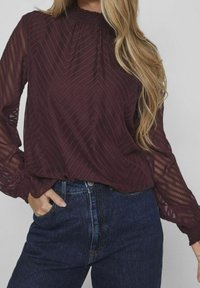 Blusa bordeaux con scollo arricciato e maniche lunghe trasparenti e a fantasia, abbinata a jeans in denim scuro a vita alta.