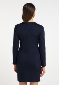 Tummansininen muotoiltu mekko, jossa on pitkähihaiset hihat, rakenteellinen kaulus ja sauma-alueet. Valmistettu joustavasta kankaasta, jossa on sileä pinta.