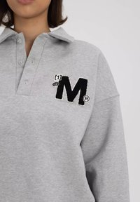 Szara, oversize polo z zapinanym kołnierzykiem, z czarnym haftowanym logo "M" oraz detalem. Miękka tekstura i casualowy design.