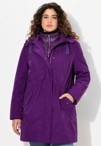 Giacca esterna waterproof viola con vita stretta, grandi tasche frontali e cappuccio rimovibile, indossata sopra una giacca interna trapuntata viola.