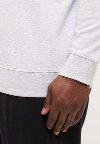 Hellgrauer Pullover mit strukturierten Rippbündchen und -saum. Nahaufnahme einer Hand, die auf dem Stoff ruht und das weiche Material sowie die Passform zeigt.