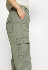 Kargo kalhoty v jasně olivově zelené barvě, s uvolněným střihem, bočními kapsami a knoflíkovými detaily na kapsách. Vyrobeno z měkké bavlněné látky.