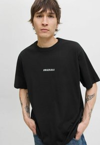 Melnas kokvilnas t-krekls ar apaļu apkakli un brīvu piegriezumu, kurā centrā ir balts uzraksts "ORIGINALS". Īsas piedurknes un minimāla tekstūra.