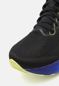 Scarpa sportiva nera con una parte superiore testurizzata, suola esterna verde brillante e accenti blu sulla intersuola. Focalizzata su prestazioni e durabilità.