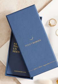 Boîte cadeau texturée bleu marine avec des lettres dorées "VIOLET HAMDEN" et un texte à l'intérieur disant "VOIR L'INVISIBLE", avec une ligne décorative en dessous.