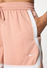 Artimas planas, kuriame matosi ranka kišenėje persikinių spalvos Nike šortų su baltais dryžiais ir raišteliu juosmenyje.