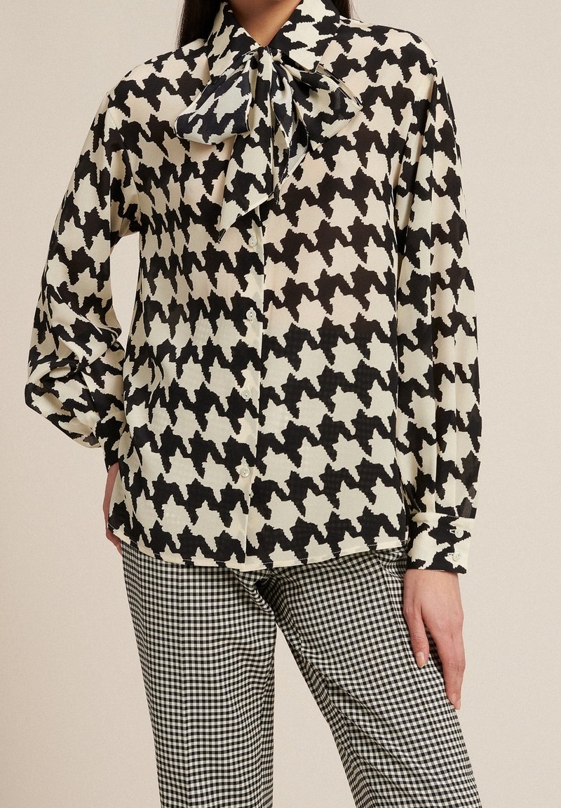 Blouse à motif pied-de-poule noir et crème avec un col lavallière, en tissu lisse, fermeture à boutons, associée à un pantalon à carreaux noir et blanc.