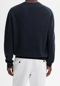 Dunkelblauer Strickpullover mit Rundhalsausschnitt, Raglanärmeln und geripptem Saum. Kombiniert mit weißen Hosen mit schwarzen Knopfakzenten.